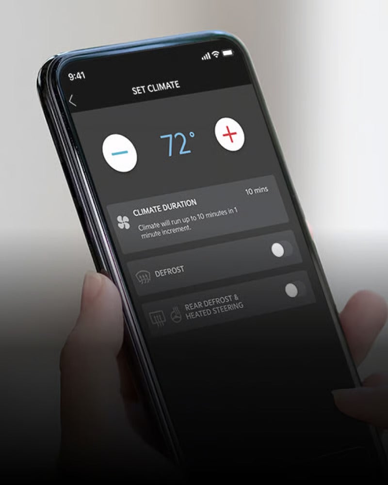 A hand holds a smartphone displaying a car climate control app set to 72 degrees, with options for defrost.