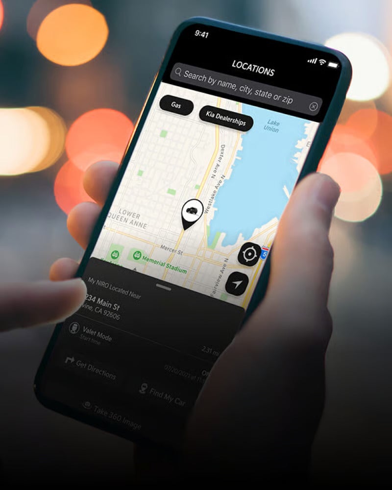 A hand holding a phone displaying a map app. The map shows Kia Dealerships and a car icon, with details like My NIRO Located Near and an address below.