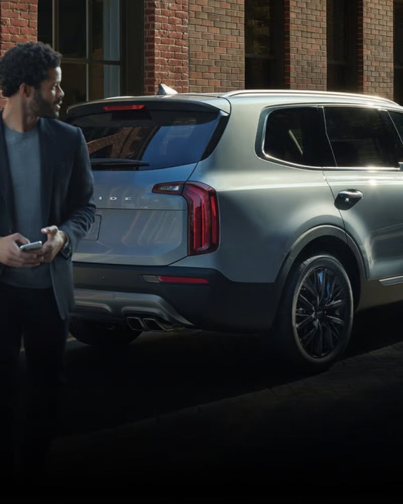 A person holding a phone looks toward a gray SUV parked in an urban setting.