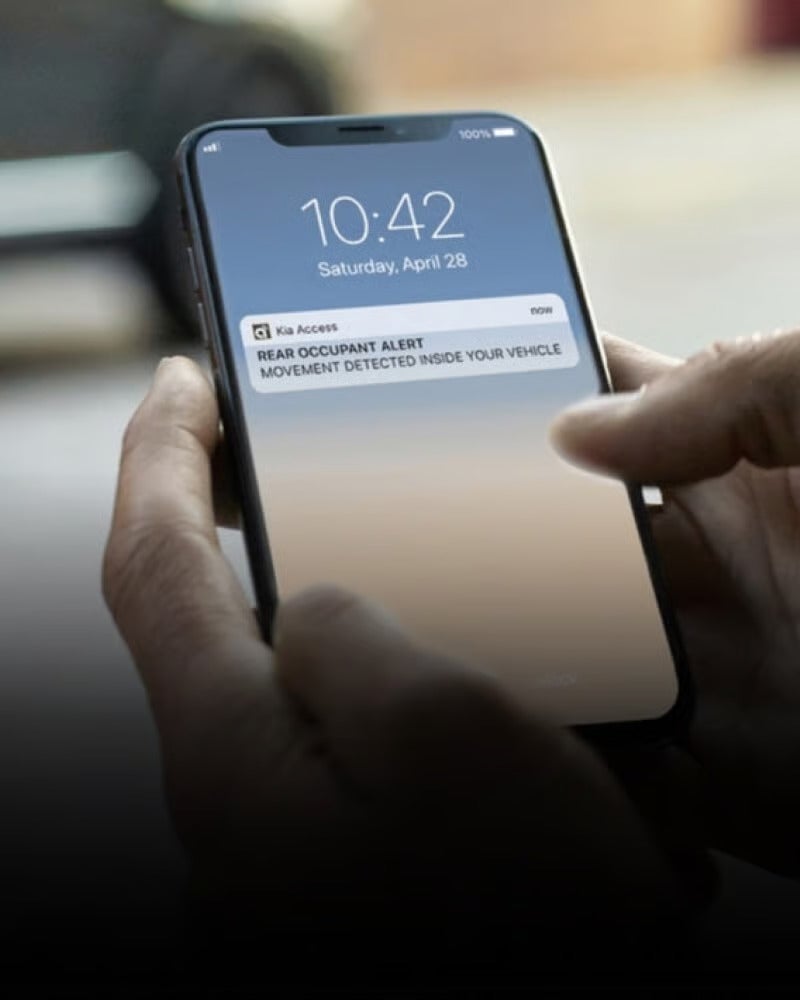 A hand holds a smartphone displaying a Rear Occupant Alert notification from Kia Access