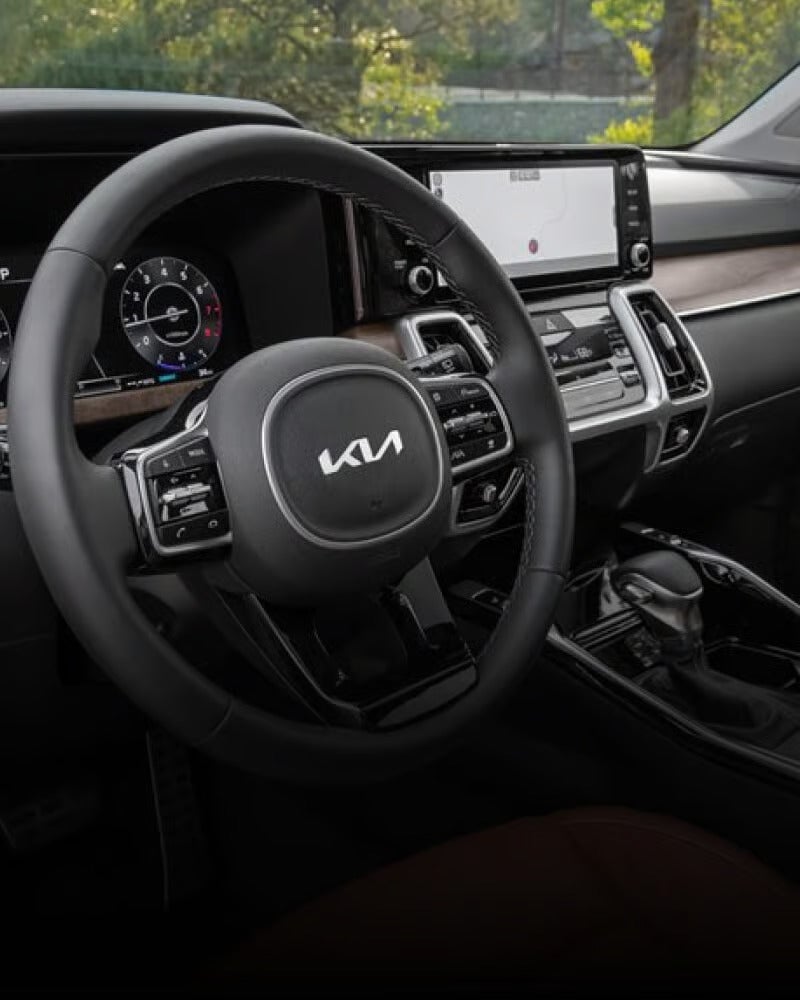 The interior of a car features a steering wheel with a Kia logo, a digital instrument cluster, and a central infotainment screen.