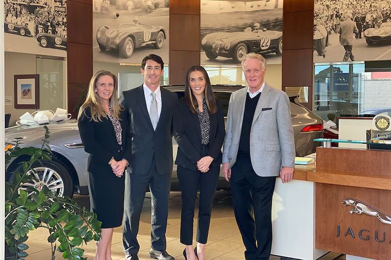 Four people in business attire stand in front of a Jaguar car inside a dealership, with vintage car photos displayed on the wall behind them.