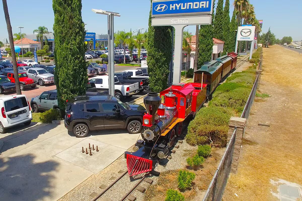A red miniature train on tracks surrounded by lush green landscaping, with a car dealership parking lot and sign in the background.