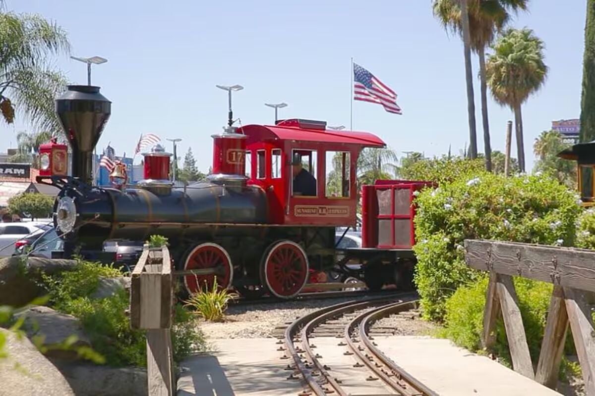 A black and red miniature train on tracks, with a U.S. flag flying behind it and palm trees in the background.